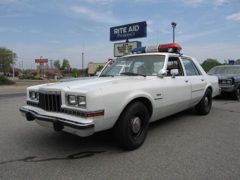 1991 Ford LTD Crown Victoria - white 1980s Dodge sedan outfitted as a police car with a roof lightbar, parked in front of a Rite Aid sign.