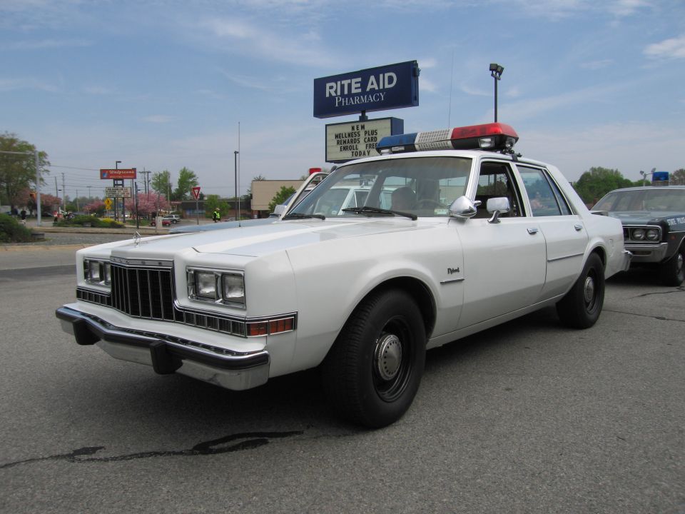 White 1980s Dodge sedan police car with roof lightbar in parking lot