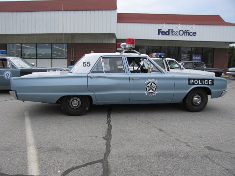 1991 Ford LTD Crown Victoria - Side view of a light blue vintage police sedan numbered 55 with a roof lightbar and star emblem on the door.