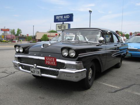1991 Ford LTD Crown Victoria - Front view of a black 1950s Ford coupe with a red State Police license plate, parked near a Rite Aid pharmacy.