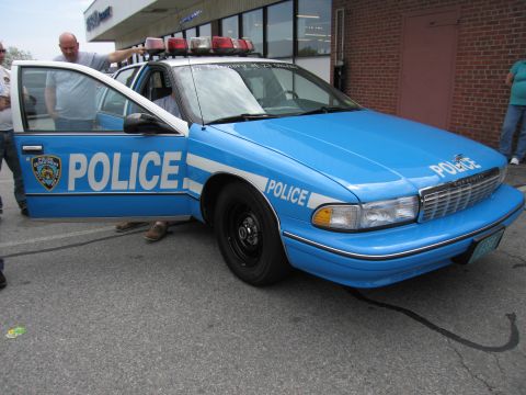 1991 Ford LTD Crown Victoria - blue NYPD-marked Chevrolet Caprice police car with its driver door open and people standing alongside in a lot.