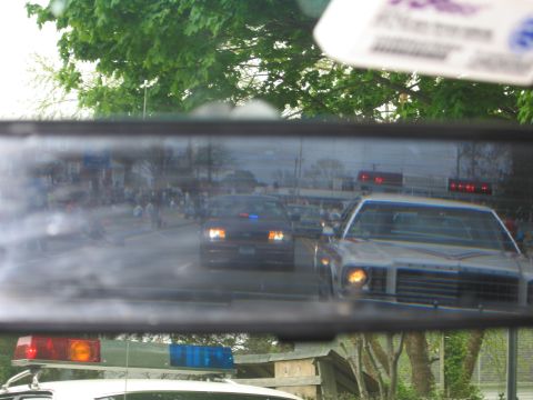 1991 Ford LTD Crown Victoria - car's rearview mirror reflects a line of police cars with emergency lights following on a street during a parade.