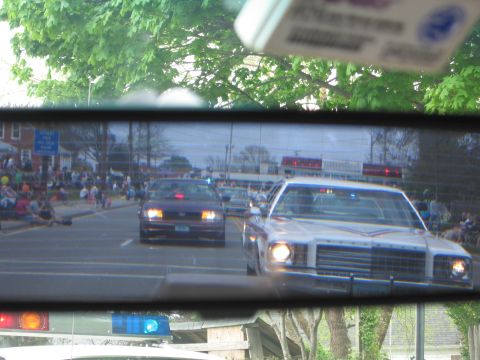 1991 Ford LTD Crown Victoria - rearview mirror reflects police cars with headlights on and a crowd of spectators lining a street during a parade.