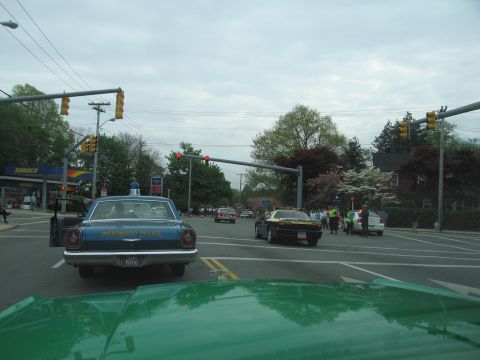 1991 Ford LTD Crown Victoria - blue vintage Westwood Police sedan waits at a red traffic light on a tree-lined town street, viewed over a green car hood.