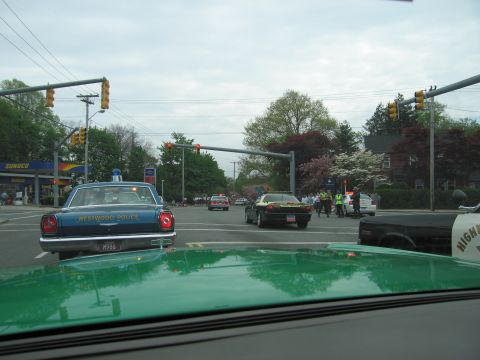 1991 Ford LTD Crown Victoria - blue Westwood Police vintage sedan sits at a town intersection near a Sunoco station, framed by a green car hood and windshield.