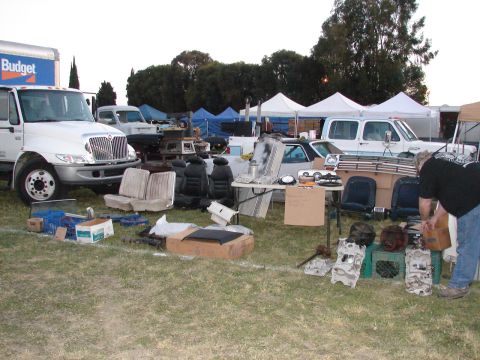 1973 Ford Ranchero - A variety of car parts are displayed on the grass at an outdoor market, with several vehicles and tents in the background.