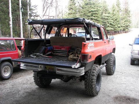 1990 Chevrolet Suburban - Modified SUV with open tailgate, parked next to other vehicles in a wooded area.
