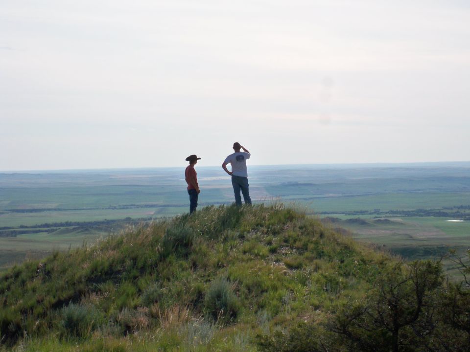 Two people stand on a grassy hill, surveying a wide, green landscape under a hazy sky