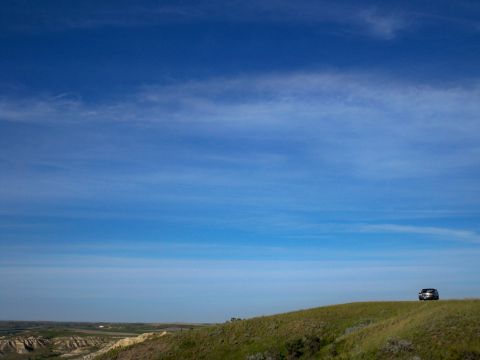 1994 Ford Bronco - A lone dark SUV sits on a grassy incline beneath a wide expanse of blue sky with wispy clouds.