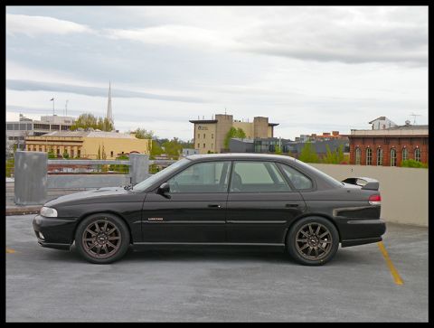 1998 Subaru Legacy - Side profile of a black 1998 Subaru Legacy sedan parked on a rooftop with city buildings in the background.