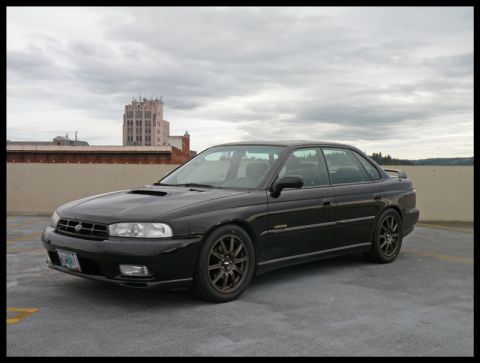 1998 Subaru Legacy - A black Subaru sedan is parked on a rooftop under a cloudy sky, with a building visible in the distance.