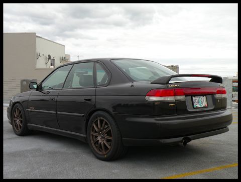 1998 Subaru Legacy - A black 1998 Subaru Legacy sedan is parked on a rooftop, a rear spoiler and aftermarket wheels.