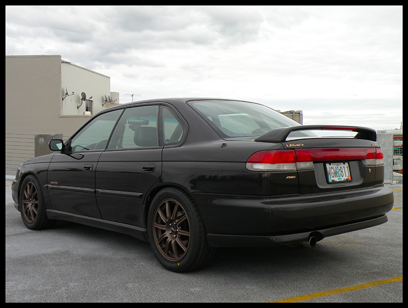 Black 1998 Subaru Legacy sedan with a rear spoiler and bronze wheels, viewed from the rear three-quarter angle