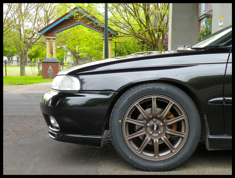 Black car's front fender and bronze BBS wheels with orange brake calipers