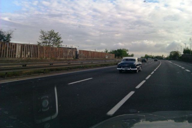 Dark blue and white 1950s sedan driving on a highway, rear view