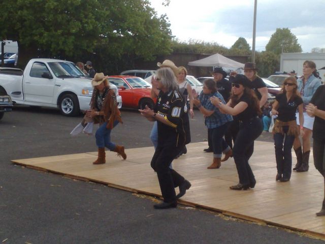 People line dancing on a wooden platform outdoors, with several cars parked in the background