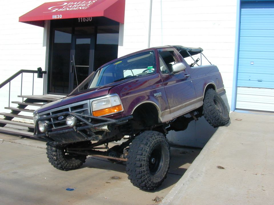 Lifted maroon topless Ford Bronco on big mud tires angled on a curb outside a business.