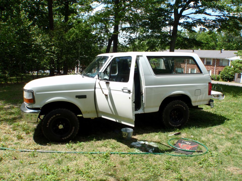 White two-door Ford Bronco SUV parked on grass, driver's door open, side view.