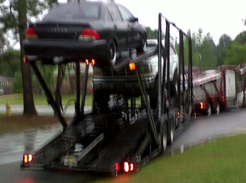 1996 Ford Bronco - A black sedan is transported on a car carrier, with another vehicle partially visible underneath.