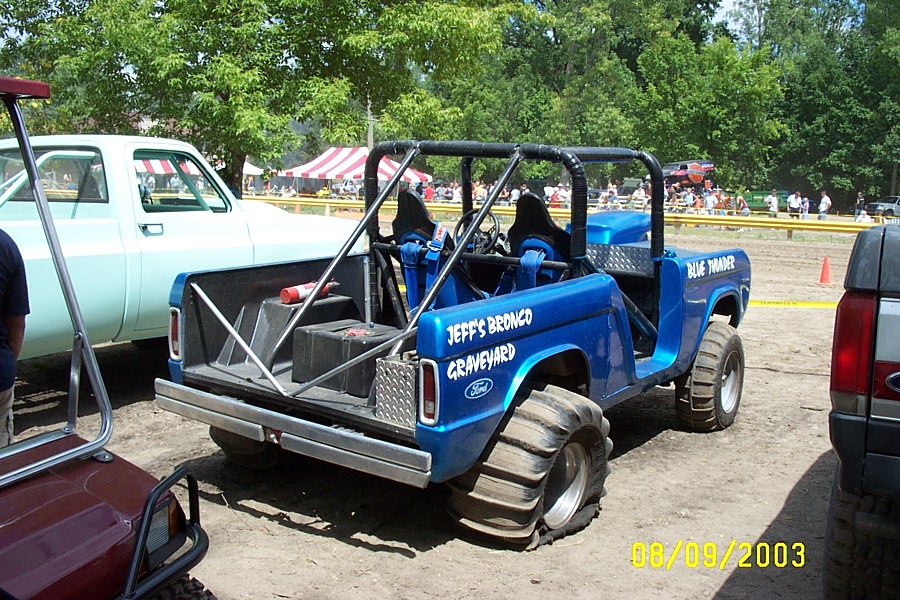 Blue off-road vehicle with roll cage and large tires, "JEFF'S BRONCO GRAVEYARD" text on side. Another light-colored truck is to the left