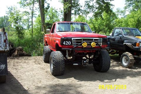 1996 Ford Bronco - A modified red pickup truck with off-road tires sits parked next to another truck in a wooded area.