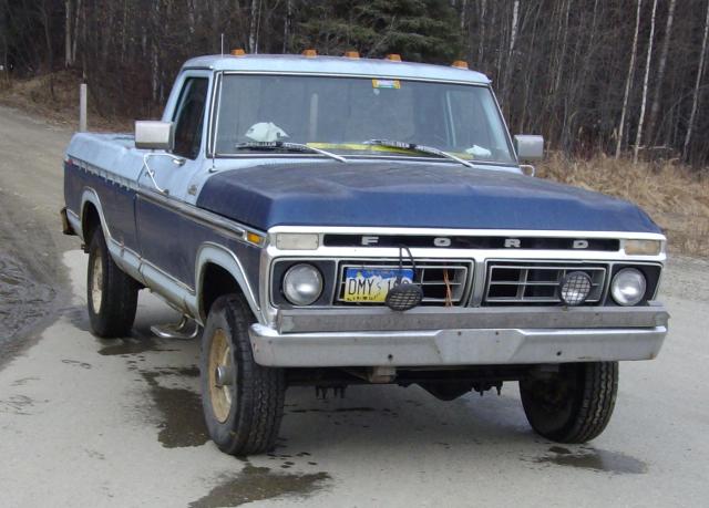 Two-tone blue and white 1970s Ford F-series 4x4 pickup on a wet gravel road