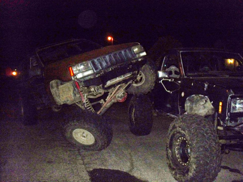 Brown SUV with lifted suspension and large tires is angled over a black SUV at night