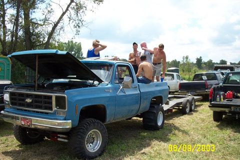 1996 Ford Bronco - A group of people are gathered around a lifted blue pickup truck with its hood open, parked next to other vehicles.