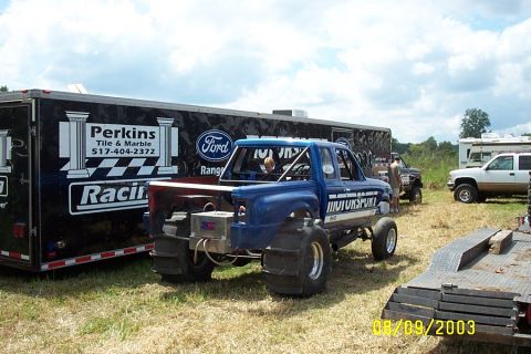 1996 Ford Bronco - A modified blue pickup truck with oversized tires and a roll cage is parked next to a trailer.