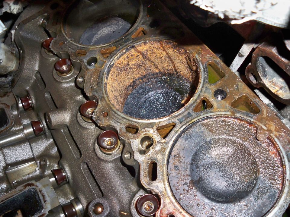 Close-up of a damaged engine cylinder with carbon buildup and rust. The cylinder head is removed, showing the piston top and surrounding metal