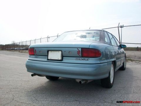 1993 Ford Taurus - The rear of a light blue Mercury Sable sedan is shown parked in an asphalt lot with a chain-link fence in the background.