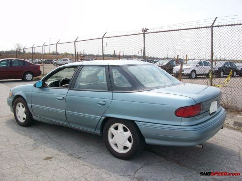 1993 Ford Taurus - This light blue sedan features a distinctive rear quarter profile and white wheels, appearing to be from the mid-1990s.