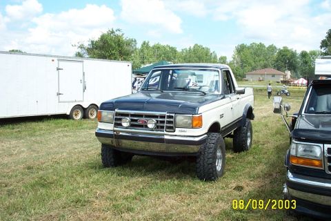 1996 Ford Bronco - A two-tone Ford Bronco with aftermarket lights sits on a grassy field next to another vehicle and a trailer.
