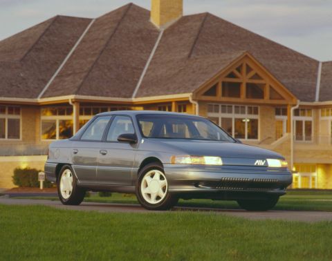 1993 Ford Taurus - A gray sedan with a distinctive grille emblem is parked in front of a large house.