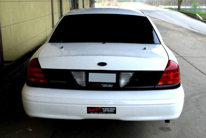 Rear view of a white sedan with tinted windows and a black accent panel between the taillights. It has a 'DARE' sticker on the bumper