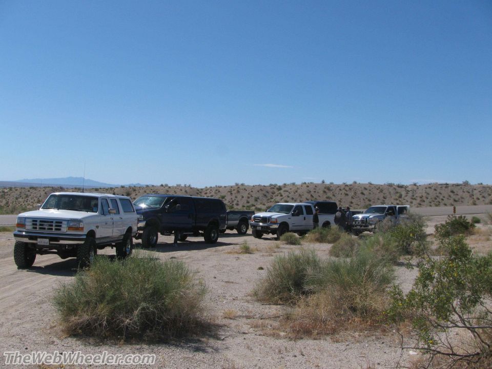 Several SUVs and trucks parked on a dirt road in a desert with scrub brush and hills