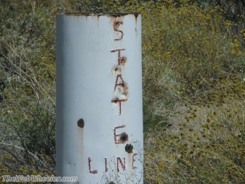 1996 Ford C-350 (Centurion Conversions) - A weathered state line marker stands in a dry, grassy field with scattered yellow wildflowers.