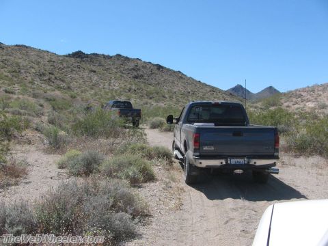 1996 Ford C-350 (Centurion Conversions) - Two pickup trucks drive on a dirt road through a desert landscape under a clear blue sky.