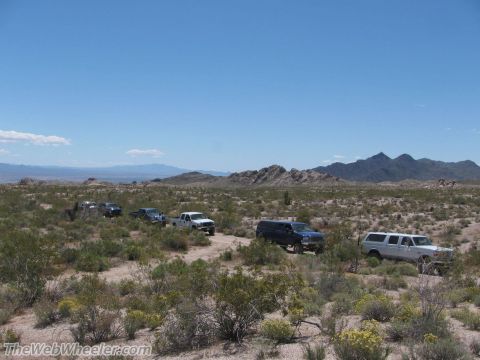 1996 Ford C-350 (Centurion Conversions) - A convoy of Ford trucks and SUVs navigates a dusty trail through a sparse desert terrain with mountains in the background.