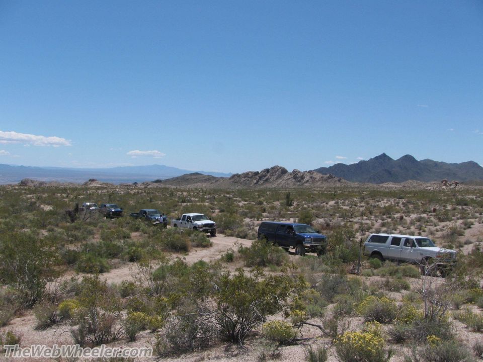 Several Ford trucks and SUVs drive on a dirt road in a desert landscape under a clear blue sky