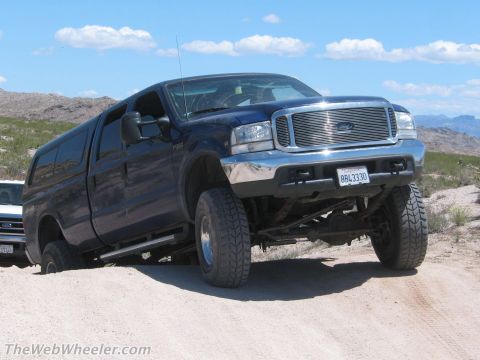 1996 Ford C-350 (Centurion Conversions) - A dark blue Ford Super Duty crew-cab pickup with a matching camper shell straddles a dirt berm, front passenger wheel drooped to show the s…