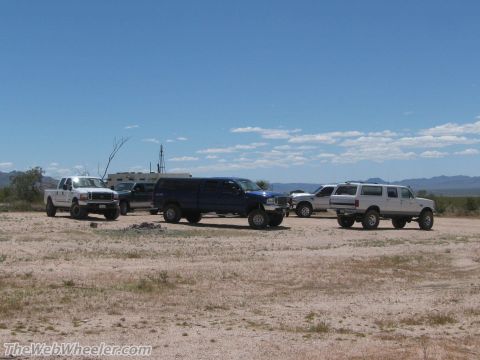1996 Ford C-350 (Centurion Conversions) - A group of modified trucks and SUVs are parked in an arid landscape with a distant building and windmill.