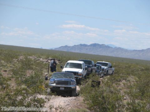 1996 Ford C-350 (Centurion Conversions) - Several Ford F-Series trucks drive on a dusty trail through a desert landscape with mountains in the background.