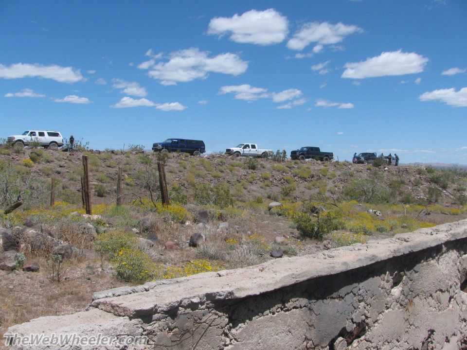 Several SUVs and trucks parked on a dry, scrub-covered hillside under a blue sky with scattered clouds