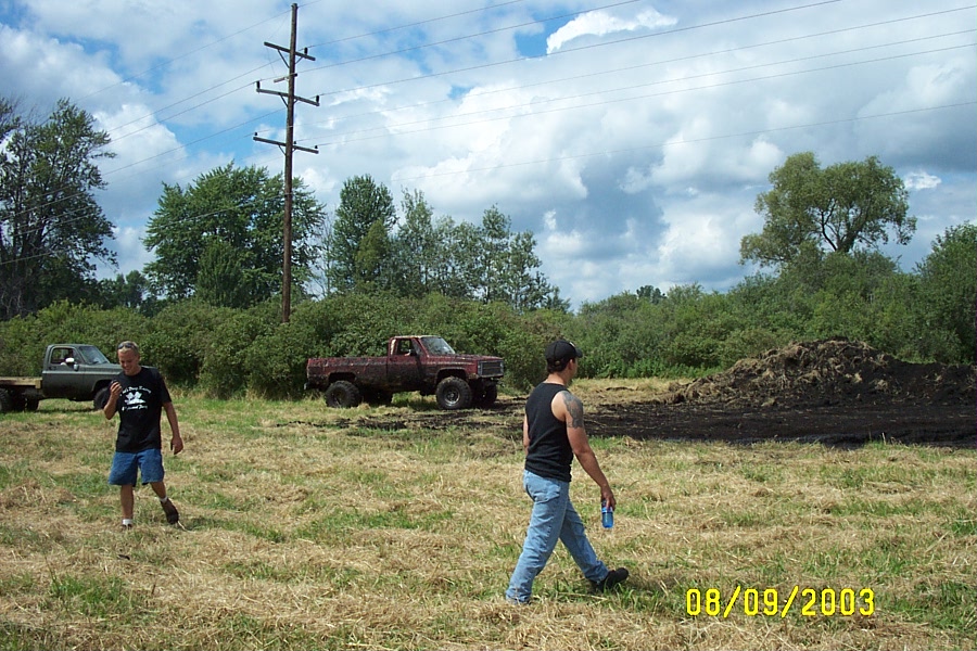 Mud-covered red square-body pickup on big tires at a mud pit with two men walking by