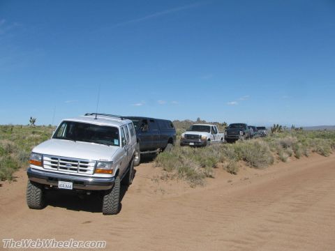 1996 Ford C-350 (Centurion Conversions) - A convoy of Ford trucks and SUVs navigate a dusty desert trail under a clear blue sky.