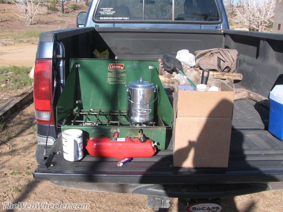 Pickup truck bed loaded with a green Coleman stove, fuel tank, pot, box, and other camping supplies