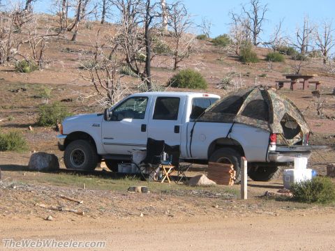 1996 Ford C-350 (Centurion Conversions) - A white Ford F-250 Super Duty pickup truck is parked in a dry, wooded area with a tent set up in its bed.