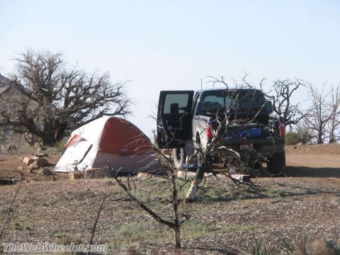 1996 Ford C-350 (Centurion Conversions) - A dark pickup truck is parked next to a tent at a campsite, with dry brush and trees in the background.