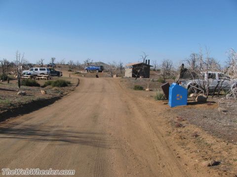 1996 Ford C-350 (Centurion Conversions) - A dirt road winds through a dry, scrubby landscape towards a campsite with several vehicles and a small building.
